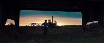 Movie still from “Out of the Furnace” (2013), directed by Scott Cooper – A person standing in a field near a factory at sunset; Extreme Wide shot, Low angle