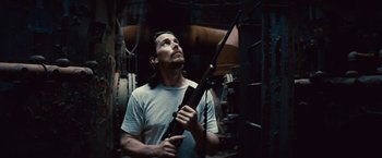 Movie still from “Out of the Furnace” (2013), directed by Scott Cooper – A man holding a rifle in a room; Medium shot, Low angle