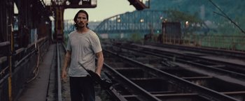 Movie still from “Out of the Furnace” (2013), directed by Scott Cooper – A man holding an umbrella while standing next to train tracks; Medium shot, Over the shoulder angle