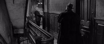 Movie still from “The Apartment” (1960), directed by Billy Wilder – A man in a suit and a man in a hat standing in front of a staircase; Medium shot, Over the shoulder angle