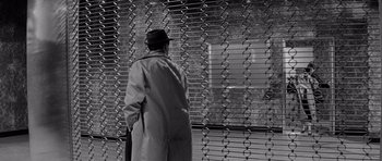 Movie still from “The Apartment” (1960), directed by Billy Wilder – A man in a trench coat and hat looking out a window; Wide shot, Over the shoulder angle