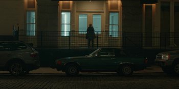 Movie still from “The Assistant” (2019), directed by Kitty Green – A car parked on the side of the road near a building; Extreme Wide shot, Low angle