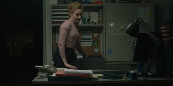 Movie still from “The Assistant” (2019), directed by Kitty Green – A woman standing at a desk in front of a laptop computer; Medium shot, Over the shoulder angle