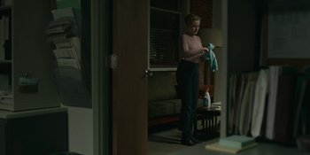 Movie still from “The Assistant” (2019), directed by Kitty Green – A woman in a pink shirt is holding a blue towel; Wide shot, Over the shoulder angle