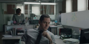 Movie still from “The Assistant” (2019), directed by Kitty Green – A man sitting at a desk in an office setting; Medium shot, Low angle