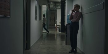 Movie still from “The Assistant” (2019), directed by Kitty Green – A woman standing in a hallway talking on a phone; Wide shot, Over the shoulder angle