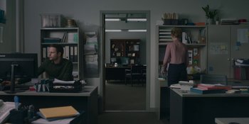 Movie still from “The Assistant” (2019), directed by Kitty Green – A person standing in a room with a lot of books on a desk; Wide shot, Over the shoulder angle