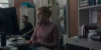 Movie still from “The Assistant” (2019), directed by Kitty Green – A woman sitting at a computer desk with a phone; Medium shot, Over the shoulder angle