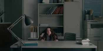 Movie still from “The Assistant” (2019), directed by Kitty Green – A girl sitting at a desk in a room; Medium shot, High angle