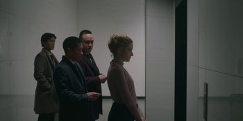Movie still from “The Assistant” (2019), directed by Kitty Green – A woman standing next to two men in a room; Medium shot, Over the shoulder angle