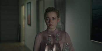 Movie still from “The Assistant” (2019), directed by Kitty Green – A woman in a pink shirt holding two bottles of wine; Medium shot, Over the shoulder angle