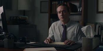 Movie still from “The Assistant” (2019), directed by Kitty Green – A man sitting at a computer keyboard in a room; Medium shot, Low angle