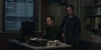 Movie still from “The Assistant” (2019), directed by Kitty Green – Two men are sitting at a desk in a dark room; Medium shot, Over the shoulder angle