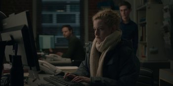 Movie still from “The Assistant” (2019), directed by Kitty Green – A woman sitting in front of a computer keyboard in front of two other men; Medium shot, Over the shoulder angle