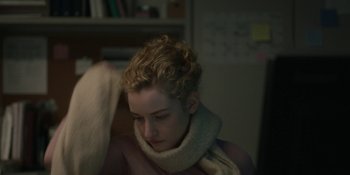 Movie still from “The Assistant” (2019), directed by Kitty Green – A young woman with curly blonde hair wearing a scarf; Close Up shot, Over the shoulder angle