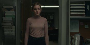 Movie still from “The Assistant” (2019), directed by Kitty Green – A woman standing in front of a bookshelf in a room; Medium shot, Over the shoulder angle