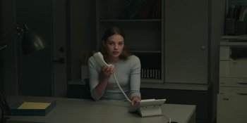 Movie still from “The Assistant” (2019), directed by Kitty Green – A woman sitting at a desk holding a phone; Medium shot, Over the shoulder angle