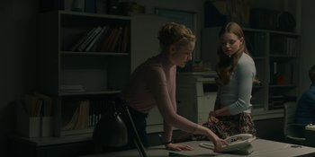 Movie still from “The Assistant” (2019), directed by Kitty Green – Two young women sitting at a table looking at a book; Medium shot, Over the shoulder angle
