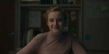 Movie still from “The Assistant” (2019), directed by Kitty Green – A woman in a pink shirt is smiling for the camera; Close Up shot, Over the shoulder angle