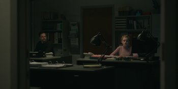 Movie still from “The Assistant” (2019), directed by Kitty Green – Two people sitting at a desk in a dark room; Wide shot, Over the shoulder angle