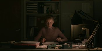 Movie still from “The Assistant” (2019), directed by Kitty Green – A woman sitting at a desk in front of papers; Medium shot, Over the shoulder angle