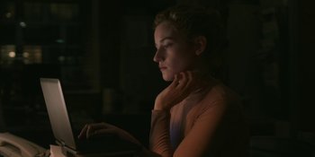 Movie still from “The Assistant” (2019), directed by Kitty Green – A woman sitting in front of a laptop computer in the dark; Medium shot, Low angle