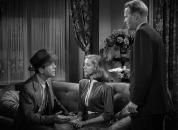 Movie still from “The Big Sleep” (1946), directed by Howard Hawks – A man and a woman sitting on a couch next to a man holding a knife; Medium shot, High angle