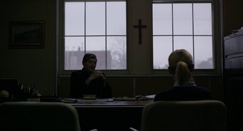 Movie still from “The Blackcoat's Daughter” (2015), directed by Oz Perkins – Two people sitting at a table in front of a cross; Medium shot, Over the shoulder angle