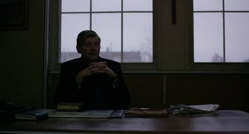 Movie still from “The Blackcoat's Daughter” (2015), directed by Oz Perkins – A man sitting at a table in front of a window; Medium shot, Over the shoulder angle