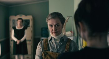 Movie still from “The Blackcoat's Daughter” (2015), directed by Oz Perkins – An older woman wearing an overalls in a room; Medium shot, Over the shoulder angle