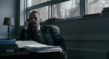Movie still from “The Blackcoat's Daughter” (2015), directed by Oz Perkins – An older man sitting at a desk talking on the phone; Medium shot, Over the shoulder angle