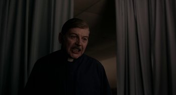 Movie still from “The Blackcoat's Daughter” (2015), directed by Oz Perkins – A man in a priest's outfit with his mouth wide open; Medium shot, Over the shoulder angle