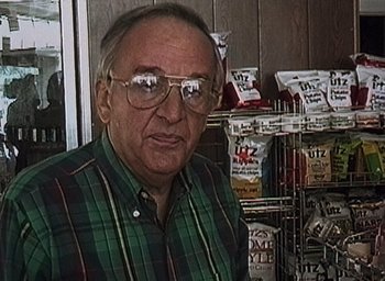 Movie still from “The Blair Witch Project” (1999), directed by Eduardo Sánchez – An older man wearing glasses standing in front of shelves of snacks; Close Up shot, High angle