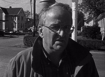 Movie still from “The Blair Witch Project” (1999), directed by Eduardo Sánchez – An older man wearing glasses standing in front of a building; Close Up shot, Over the shoulder angle