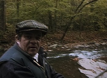 Movie still from “The Blair Witch Project” (1999), directed by Eduardo Sánchez – An older man in a hat and jacket standing by a river; Medium shot, High angle