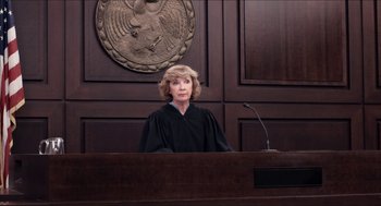 Movie still from “The Bling Ring” (2013), directed by Sofia Coppola – A judge in a courtroom with a microphone in front of him; Medium shot, Over the shoulder angle