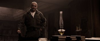 Movie still from “The Book of Eli” (2010), directed by Allen Hughes – A man standing in front of a table with a glass lamp on top of it; Medium shot, Low angle
