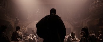 Movie still from “The Book of Eli” (2010), directed by Allen Hughes – A man standing in front of an audience in a dark room; Wide shot, Over the shoulder angle
