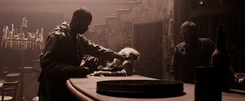 Movie still from “The Book of Eli” (2010), directed by Allen Hughes – A man sitting at a table with a cat on his lap; Medium shot, Over the shoulder angle
