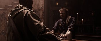 Movie still from “The Book of Eli” (2010), directed by Allen Hughes – A man sitting at a table with another man in front of him; Medium shot, Over the shoulder angle