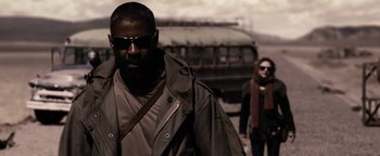 Movie still from “The Book of Eli” (2010), directed by Allen Hughes – A man with a beard and sunglasses walking next to a woman in front of a bus; Medium shot, Low angle