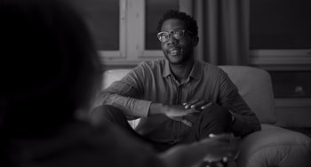 Movie still from “Paris, 13th District” (2021), directed by Jacques Audiard – A black and white photo of a man sitting on a couch; Close Up shot, Over the shoulder angle