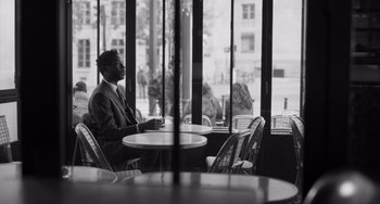 Movie still from “Paris, 13th District” (2021), directed by Jacques Audiard – A black and white photo of a man sitting at a table in a restaurant; Medium shot, Over the shoulder angle