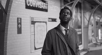 Movie still from “Paris, 13th District” (2021), directed by Jacques Audiard – A black and white photo of a man standing in front of a sign; Medium shot, Low angle