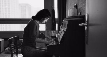 Movie still from “Paris, 13th District” (2021), directed by Jacques Audiard – A woman sitting in front of an upright piano in a room; Medium shot, Low angle