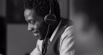 Movie still from “Paris, 13th District” (2021), directed by Jacques Audiard – A man wearing headphones and a vest smiling for the camera; Close Up shot, Over the shoulder angle