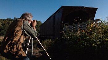 Movie still from “The Bridges of Madison County” (1995), directed by Clint Eastwood – An older man sitting in a chair looking at a covered bridge; Wide shot, Low angle