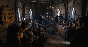 Movie still from “The Brothers Grimm” (2005), directed by Terry Gilliam – A group of people sitting in a room with a cross; Wide shot, High angle