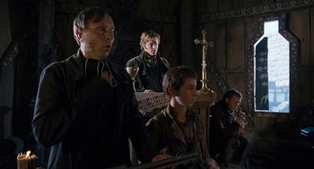 Movie still from “The Brothers Grimm” (2005), directed by Terry Gilliam – A group of people sitting in a room with a cross in the background; Medium shot, Low angle