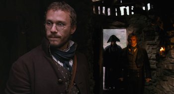 Movie still from “The Brothers Grimm” (2005), directed by Terry Gilliam – A man with a beard and glasses standing next to another man in a hat; Medium shot, Over the shoulder angle